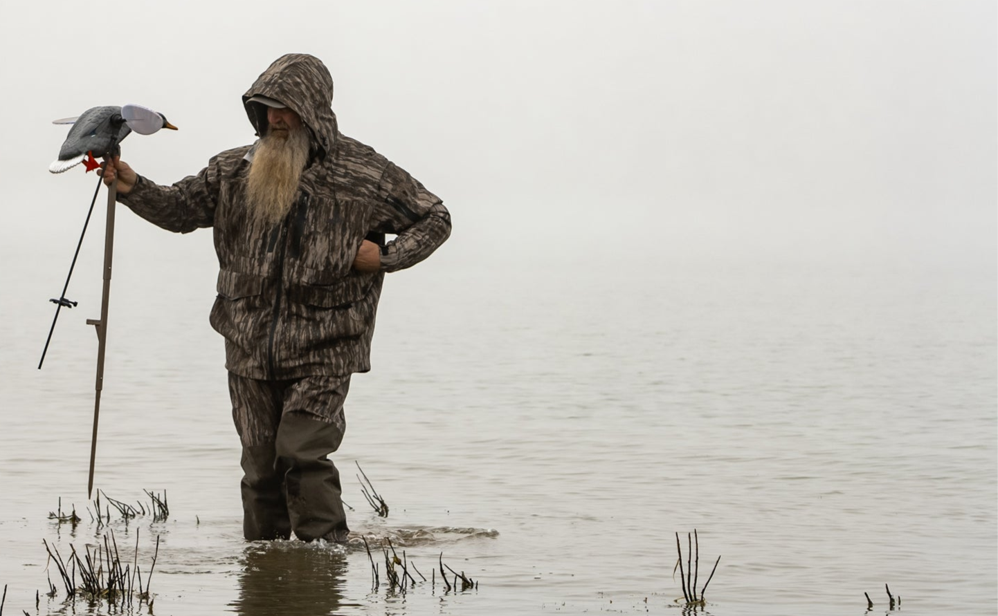 Person in camouflage wading through water holding a bird and a device, with a misty background.